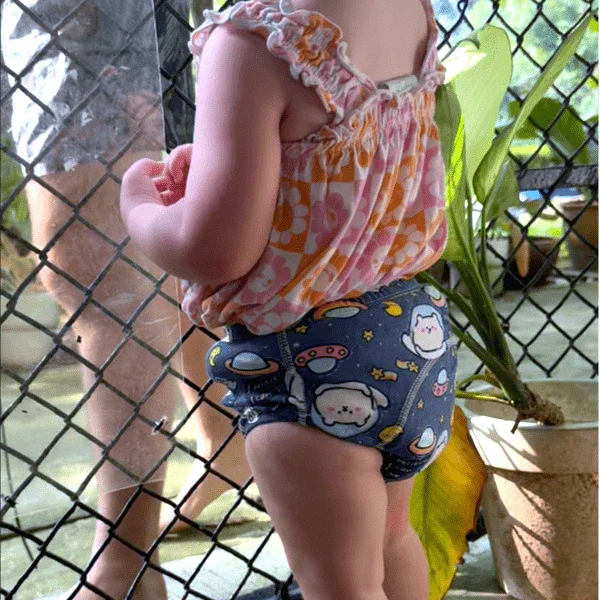 Małe dziecko w kolorowym body z motywem kotów i planet, stoi przy ogrodzeniu